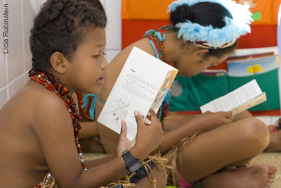 Menino e menina indígenas, sentados no chão em sala de aula, lêem cartilhas